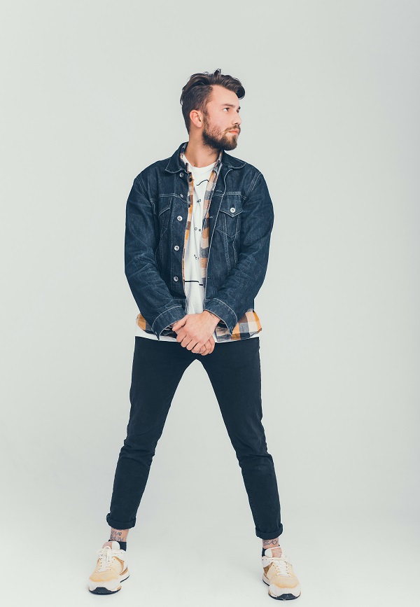 A person wearing a denim jacket, a striped shirt, black pants, and sneakers, standing against a light background. 
